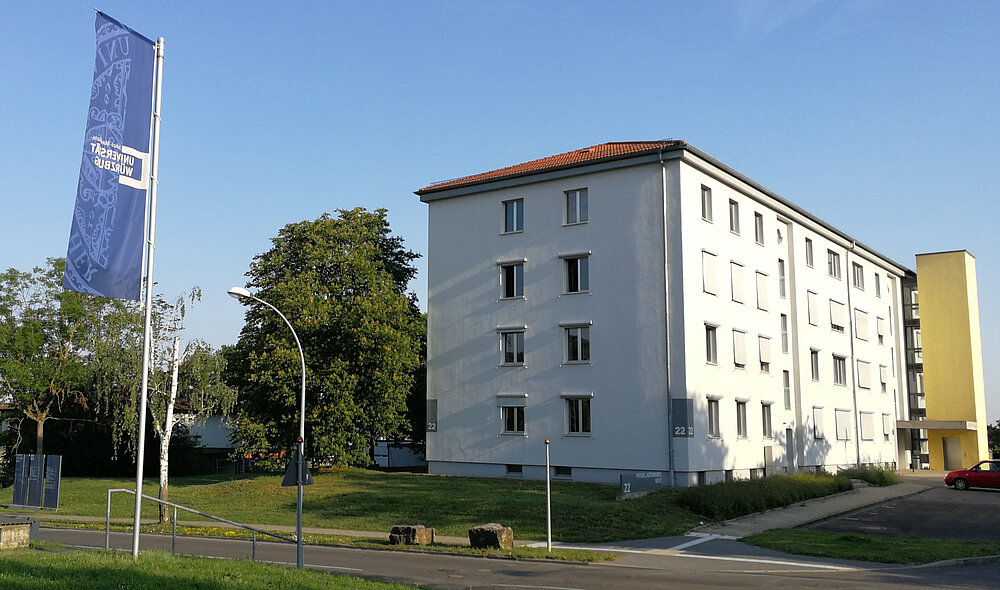von links blau-weiße Flagge der Uni WÜ, weißes Gebäude mit gelbem Vorbau im Hintergrund (Aufzug)