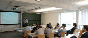 lecture hall: Speaker stands in the background at the board. About 10 persons sit in front of him with their backs at the camera.