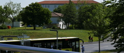Vordergrund: Bus-Haltestelle "Am Hubland" mit stehendem Bus. Hintergrund: Wiese, Stabsstelle Sicherheit, Bäume, Geb. 22.