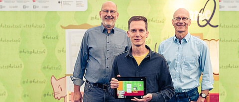 Die beiden Sprecher des Exzellenzclusters ct.qmat, Ralph Claessen (l.) und Matthias Vojta (r.), präsentieren mit dem App-Designer Philipp Stollenmayer ihr gemeinsames Projekt, die Spiele-App „Katze Q“.